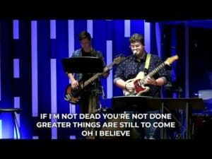 A man is playing a guitar in front of a stage with the words, if you're dead you're done greater things.