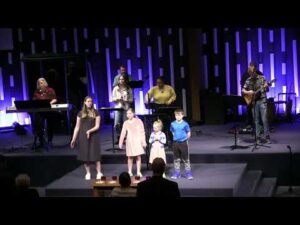 A group of children and adults perform on a stage with musical instruments and microphones, with a blue and purple lighted backdrop.
