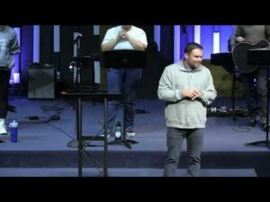 A man stands and speaks on stage while others stand behind him with musical instruments and microphones in a church or auditorium setting.
