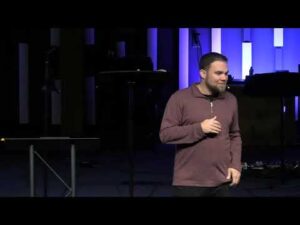 A man in a maroon sweater stands on a stage speaking, with vertical blue and white lights and equipment visible in the background.