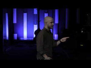 A bald man in a dark shirt stands indoors, pointing to his right, with a blue-lit background featuring vertical light panels and a musical instrument.