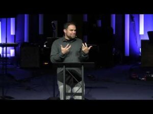 A man stands behind a podium, gesturing with his hands while speaking on a dimly lit stage with blue vertical lights in the background.