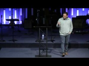 A man stands and gestures while speaking on stage in front of a podium, with musical equipment and blue-lit panels in the background.