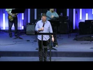 A man stands at a podium speaking into a microphone on a stage, with musicians and instruments in the background.