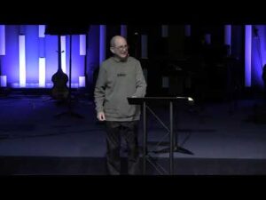 A man in a gray hoodie stands at a podium on a stage with blue vertical lights and musical instruments in the background.