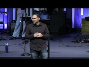 A man stands and speaks on stage in front of a microphone, with musical equipment and purple lighting in the background.