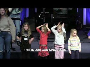 Four young children stand on stage with adults, some with hands on their heads, in front of musical equipment. Text at the bottom reads, "THIS IS OUR GOD KING JESUS.