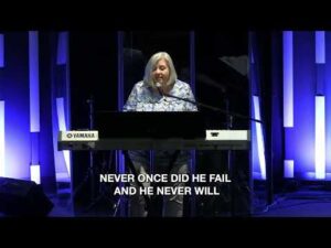A person plays a Yamaha keyboard and sings on stage with blue vertical lights in the background; subtitles read, "NEVER ONCE DID HE FAIL AND HE NEVER WILL.