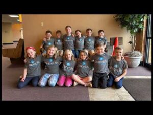 A group of thirteen children wearing matching "GraceLife" t-shirts pose together indoors, some sitting on the floor and others kneeling behind them, smiling at the camera.