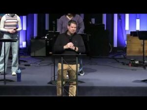 A man stands at a podium speaking on a stage, with musical equipment and two other people visible in the background.
