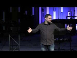 A man stands on a stage with his arms open, facing the audience. Behind him are musical instruments and vertical light panels.