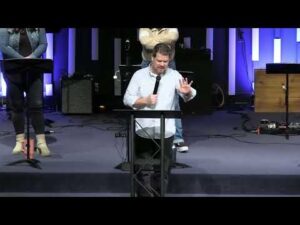 A man stands behind a podium speaking into a microphone on a stage, with people and musical equipment visible in the background.