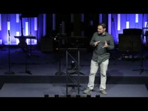 A man stands on a dimly lit stage with musical instruments in the background, speaking and gesturing with both hands.