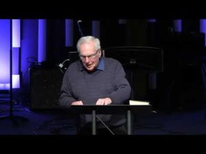 An older man with gray hair and glasses speaks at a podium on a dimly lit stage with blue and black background elements.