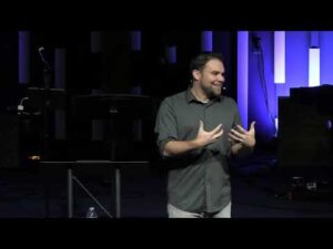 A man stands on a dimly lit stage, gesturing with his hands while speaking. There is a music stand and microphone in the background.
