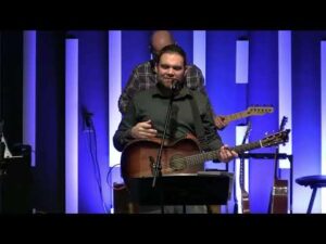 A man stands on stage holding an acoustic guitar and speaking into a microphone, with another musician and several guitars visible in the background against a blue-lit backdrop.
