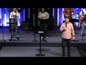 A group of people stand on a stage with microphones and musical instruments; one man is speaking at the front while others prepare behind him.