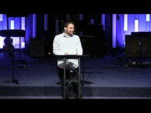 A man stands at a podium on a stage, speaking in front of a backdrop with vertical light panels and musical equipment.