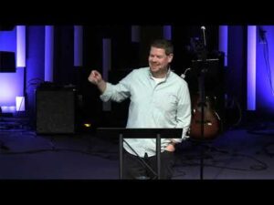 A man stands at a podium speaking on a stage with musical instruments and equipment in the background, under blue and purple lighting.