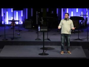 A man stands on a stage next to a small table with a water bottle, speaking to an audience; musical instruments and equipment are visible in the background.