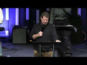 A man stands at a podium holding a microphone and reading, with another person standing behind him on a stage with blue and white vertical lighting.