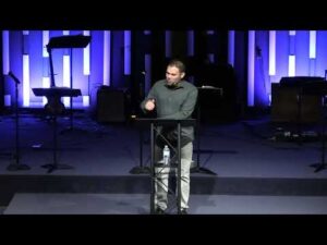A man stands at a podium speaking in a dimly lit auditorium with musical instruments in the background and blue lighting on the wall.