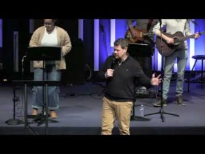 A man speaks into a microphone on stage while others stand behind him playing music; music stands and instruments are visible.