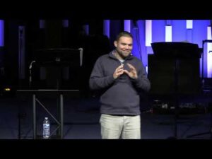 A man stands on a stage with a water bottle on a stand beside him, speaking to an audience with purple and blue lights in the background.