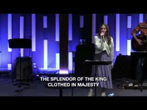 A young woman sings on a stage with blue vertical lights behind her. Song lyrics "THE SPLENDOR OF THE KING CLOTHED IN MAJESTY" are displayed at the bottom.