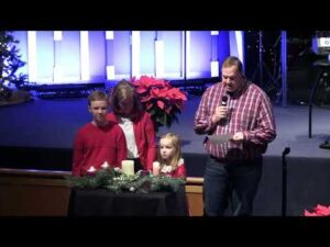 A man reads from a paper with a microphone while standing with a woman, a boy, and a girl by a table with lit candles and holiday decorations in a church setting.