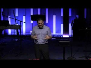 A man stands on a dimly lit stage with musical instruments in the background, gesturing with both hands while speaking.