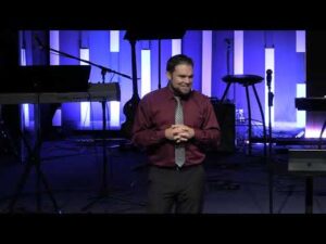 A man in a maroon shirt and tie stands on a stage with musical instruments and blue vertical lights in the background.