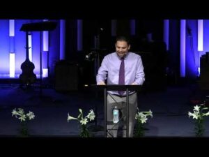A man in a light purple shirt and tie speaks at a podium on a stage decorated with white flowers, with musical instruments and equipment in the background.