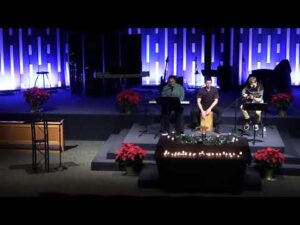 Three people perform music on a dimly lit church stage decorated with poinsettias and candles, with purple and blue lighting in the background.
