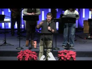 A man speaks at a podium in a church with poinsettias in front and musicians standing behind him on stage.