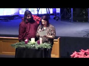 Two people stand at a table with candles and greenery; one reads from a paper while the other looks on in a church setting.