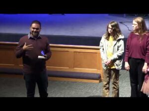 A man speaks while holding a book, standing next to two people on a stage with wooden paneling and a blue-lit background.