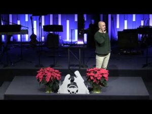 A man stands on a stage beside a nativity scene and red poinsettias, with musical instruments and blue backlighting in the background.