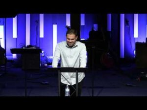 A man stands at a podium speaking in front of vertical blue and white light panels on a stage with musical equipment in the background.
