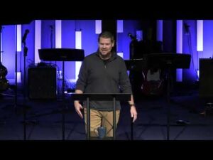 A man stands behind a podium, speaking on a stage with musical instruments and equipment in the background, lit by vertical blue lights.