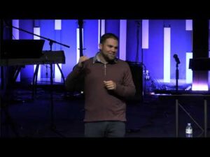 A man stands on a dimly lit stage in front of vertical light panels, gesturing with one hand. Musical equipment and a microphone are visible in the background.