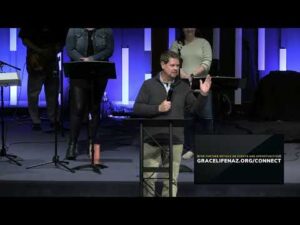 A man speaks at a podium on a stage with a microphone, while others stand behind him. Text on screen reads “GRACELIFENAZ.ORG/CONNECT.”.
