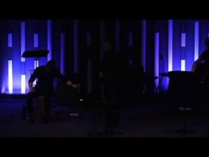 Two people on a dimly lit stage with vertical blue light panels in the background; music stands and equipment are visible.