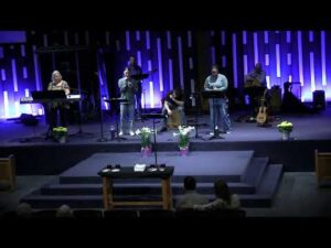 A band performs on stage in a church with blue lighting, surrounded by musical instruments and potted flowers.