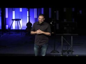 A man stands on a stage in front of blue-lit vertical panels, with a stool, guitar, and microphone stand visible in the background.