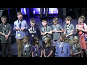 A group of children stand on stage wearing medals around their necks; some are looking at the camera, while others look elsewhere.