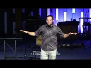 A man stands on a stage with arms outstretched, speaking to an audience. Musical instruments and equipment are visible behind him. Subtitles read: "THE ASSOCIATION IS THE MISSION OF THE CHURCH!.