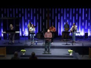 A group of musicians and a speaker stand on a church stage, lit by blue vertical lights, with musical instruments and microphones visible.