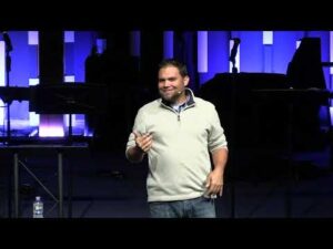 A man in a light sweater stands on a stage holding a microphone, with blue and purple lights and musical equipment in the background.