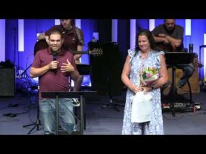 A man speaks at a podium while a woman in a floral dress stands beside him holding a bouquet of flowers; musicians sit in the background on stage.